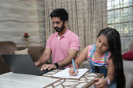 Indian father doing office work on laptop while her little daughter doing school home work. Concept of father and daughter working from home