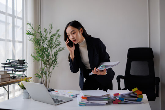 Asian businesswoman managing a heavy workload, talking on a smartphone while holding papers, surrounded by stacks of documents and using a laptop in a modern office