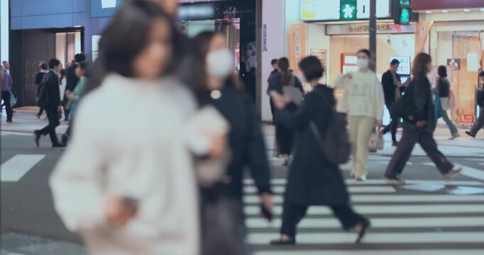 Unrecognisable people crowd crossing zebra crossing on busy urban street at night in modern city. Blurred foreground figures crossing road, motion in rush hour at Tokyo downtown. Crowded avenue 