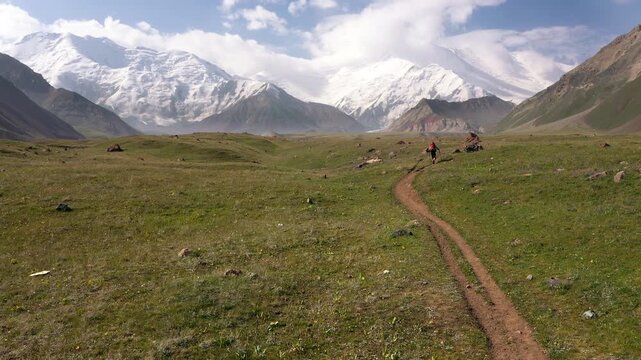 Lonely climber starting ascending route toward snow-capped Lenin peak in Pamir mountains from basecamp, alpine hiking adventure in Kyrgyzstan wilderness, high altitude expedition landscape, 4K
