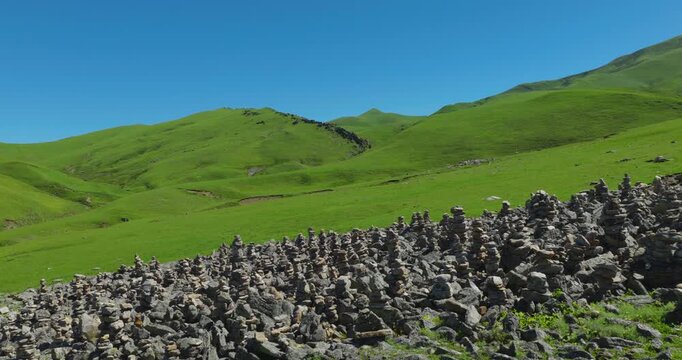 Badimalika region, Nepal, aerial shot of a vast field of stacked stone cairns on a green hillside in Triveni Patan, revealing rolling alpine pastures under a bright blue sky.