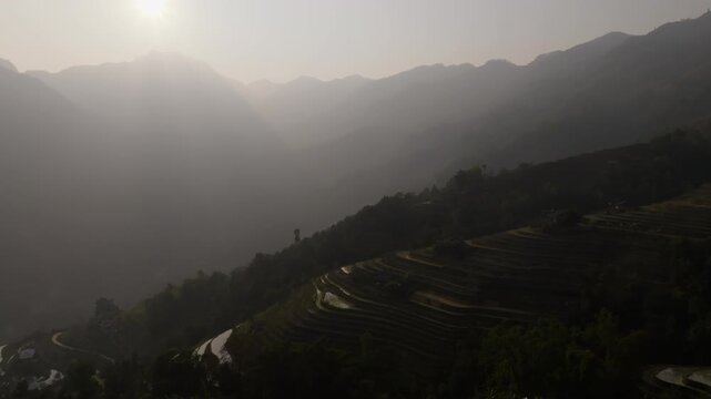 Aerial view of a hazy mountain valley at sunset with a white-roofed house and terraces catching light on a dark slope in Ho&agrave;ng Su Ph&igrave;, Vietnam, backlit dusk.