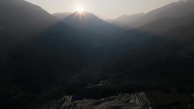 Drone glides toward a sun setting behind a mountain peak in Ho&agrave;ng Su Ph&igrave;, Vietnam; hazy ridgelines fade in the distance while terraces catch light at the bottom.