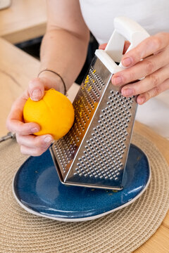 Culinary process. Zest grating. Female chef hand making aroma fruit ingredient from fresh orange using metal grater on kitchen table.