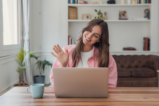 Woman smiling, making a greeting gesture with her hand during an online video call on a laptop, engaging in remote work or virtual communication from a home office setup