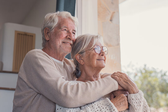 Elderly couple enjoys happy retirement while standing by a bright window at home. White-haired husband embraces his wife from behind as they look outside, representing aging, freedom and companionship