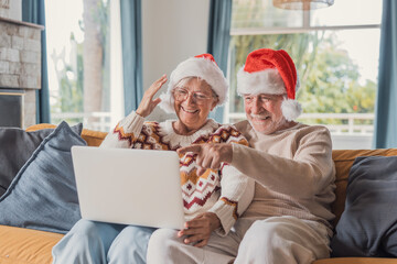 Senior couple wave and point at a laptop during a Christmas video call at home. Wearing Santa hats on the sofa, they share joyful festive greetings from a warm, bright living room with family online. © Daniel