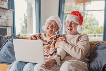 Senior couple laugh together during a Christmas video call on a laptop in their bright living room. Elderly man holds his wife close on the sofa, sharing joyful holiday greetings with family online. © Daniel