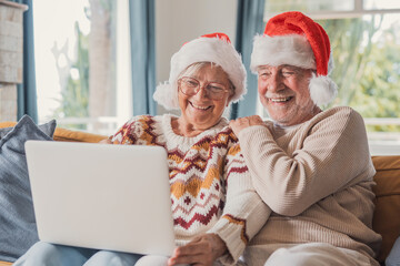 Senior couple laughs during a Christmas video call on a laptop while sitting on a sofa in their living room. Elderly woman and bearded man in Santa hats share joyful festive greetings with family. © Daniel