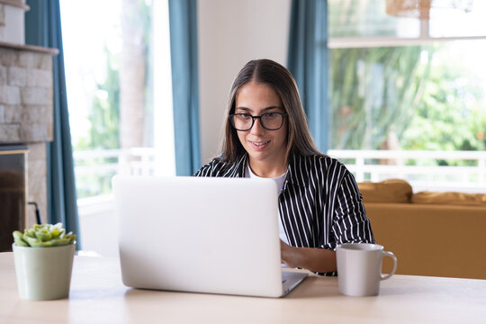 Woman creates a blogging post on her laptop at home, smiling while she writes. A mug and plant accompany content creation, blog writing, digital publishing and freelance creative work.
