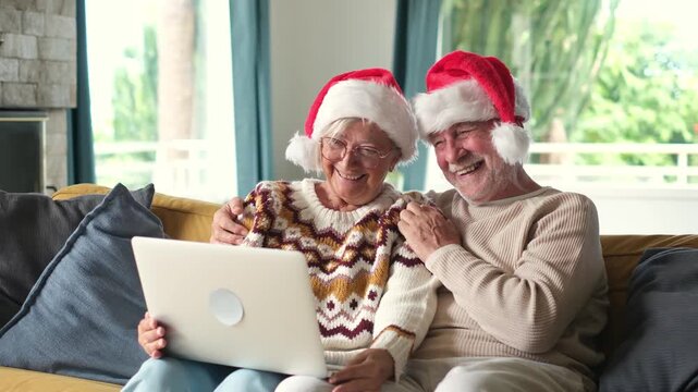 Cute elderly couple wearing santa hats and sweaters sitting on the sofa at home while video calling family on a laptop, smiling and enjoying christmas holidays together with their loved ones