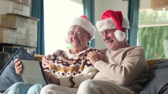 Happy old couple wearing santa hats video calling their family on a tablet computer, smiling and waving during the christmas holidays while sitting comfortably on the sofa at home