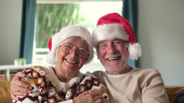 Senior couple laugh warmly while embracing on a sofa in a bright living room. Elderly man holds his wife as they wear Santa hats, look at the camera and celebrate Christmas with festive holiday joy.