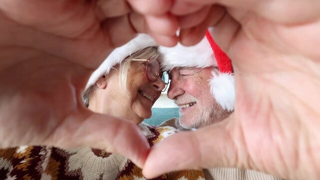 Cute senior couple making a heart shape with their hands while looking at the camera. Happy elderly husband and wife wearing santa hats, smiling together, and showing a love gesture