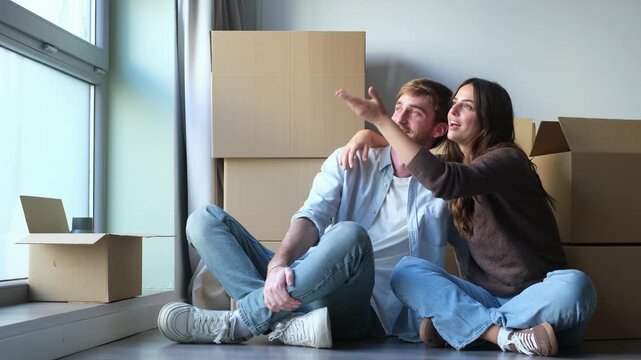Couple sits on the floor, talks and points toward the window while planning their new apartment. Moving boxes and daylight frame an affectionate conversation about arranging their new home together.