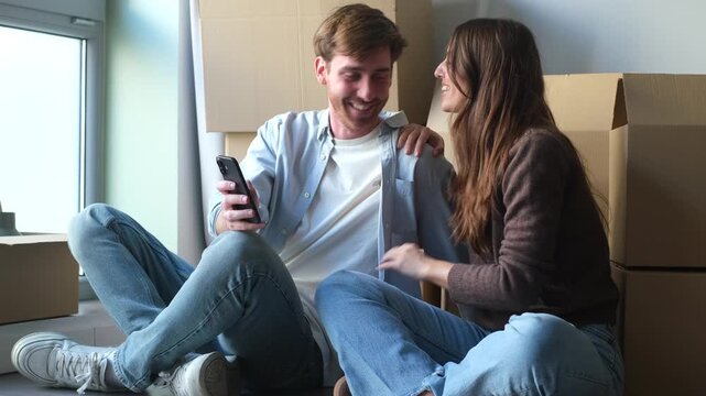 Excited young couple sitting on the floor of their new apartment, laughing while browsing on a smartphone, surrounded by cardboard moving boxes. They are planning their future together