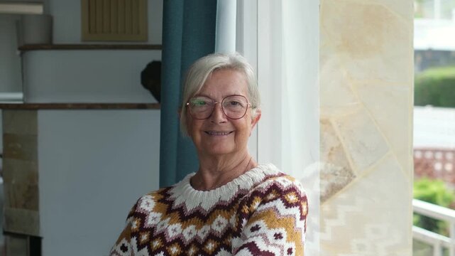 Senior woman with silver hair smiles directly at the camera beside a bright window. Her crossed-arm pose presents relaxed retirement, independent living, healthy aging, home comfort and wellness.