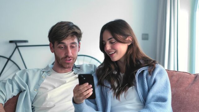 Cheerful boyfriend and girlfriend sitting together on the sofa at home while using a mobile phone, scrolling social media, shopping online, or watching videos and talking about it
