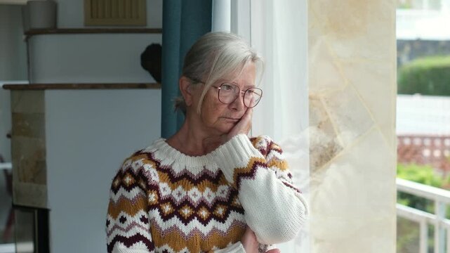 Thoughtful elderly woman with white hair and glasses looking sadly out the window, feeling lonely and isolated at home. Mature person suffering from depression and melancholy during retirement