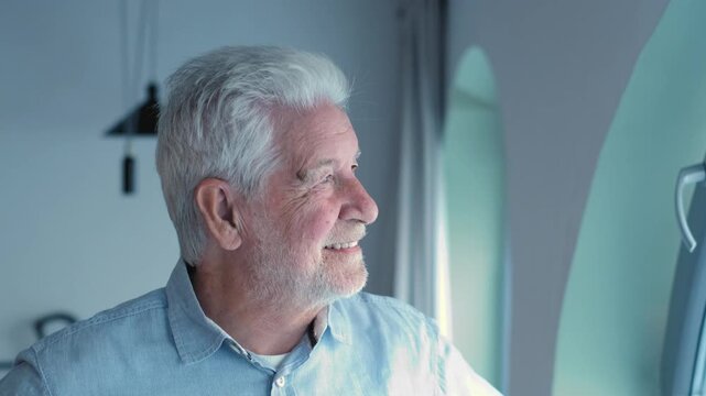 Senior man looks toward the window, then turns to smile at the camera in daylight. His changing expression conveys retirement, healthy aging, independent living, home comfort and wellbeing.