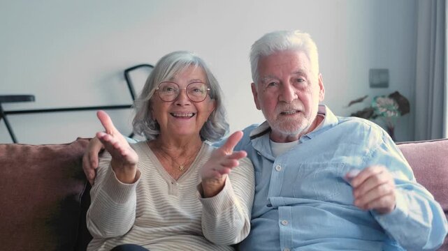 Senior caucasian couple sitting on a sofa using technology for a video call with family, waving and smiling at the camera in their living room, enjoying a happy retirement together