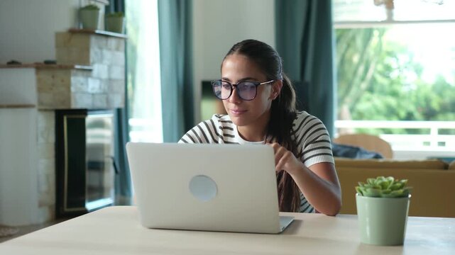 Beautiful young female freelancer wearing glasses and concentrating on her work, typing on a laptop computer keyboard while sitting at a desk in her cozy, light-filled living room