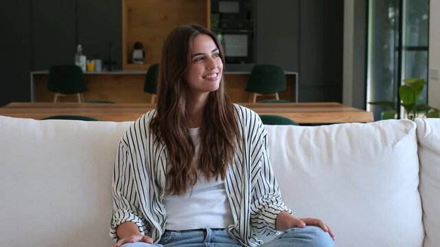 Content young woman with long hair smiling peacefully while resting on a comfortable sofa in her modern living room, enjoying a quiet moment of happiness and tranquility at home