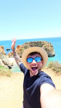 Young smiling influencer with sunglasses and a hat filming himself while spinning and showing the beautiful ocean view with rock formations during a sunny summer day in lagos, algarve, portugal