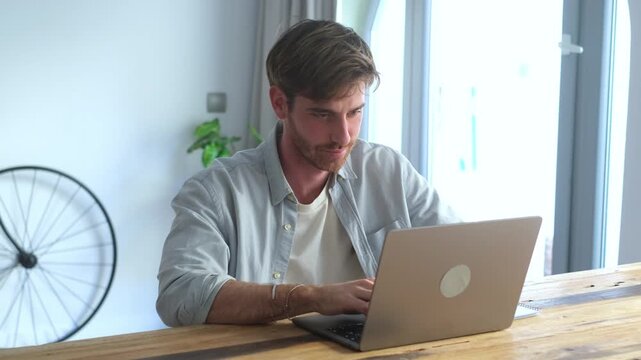 Young handsome freelancer working from home, typing on his laptop and writing down notes while studying online, focused on his tasks in a modern and bright apartment living room