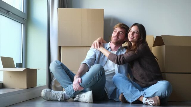 Couple sits among cardboard boxes, points across their new apartment and shares a joyful hug. Their animated conversation expresses excitement, affection and dreams of settling into a shared home.