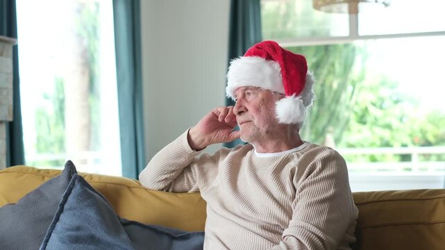 Pensive and melancholic elderly man sitting alone at home on the sofa during christmas holidays, wearing a santa hat and looking out the window with a thoughtful and sad expression