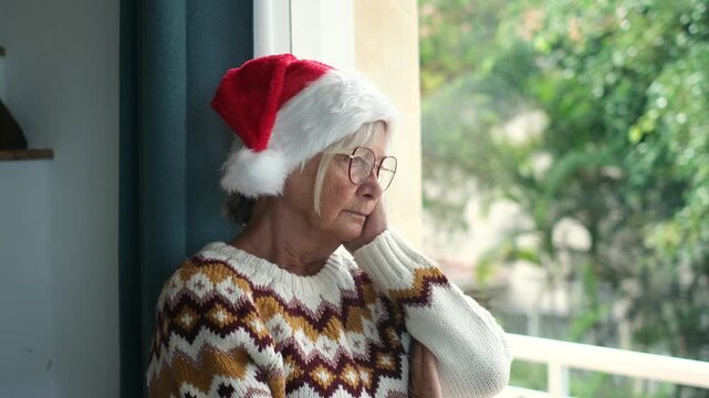 Elderly woman spends a quiet Christmas morning leaning on the window with her cheek in hand. Her solemn gaze toward greenery conveys solitary reflection, sadness and holiday isolation at home.