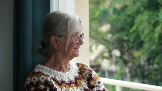 Senior woman leans near a window and smiles gently at the trees outside. Her peaceful side profile in daylight represents retirement, independent living, wellness, relaxation and aging at home.