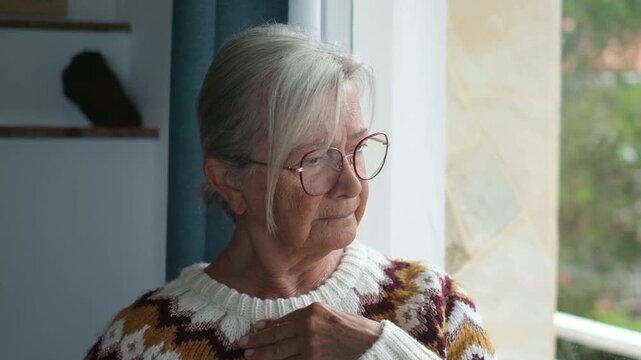Senior woman stands at a bright window and moves her hand from neck to chin in thought. Her pensive gaze outside supports themes of retirement, aging, independent living, reflection and wellness.