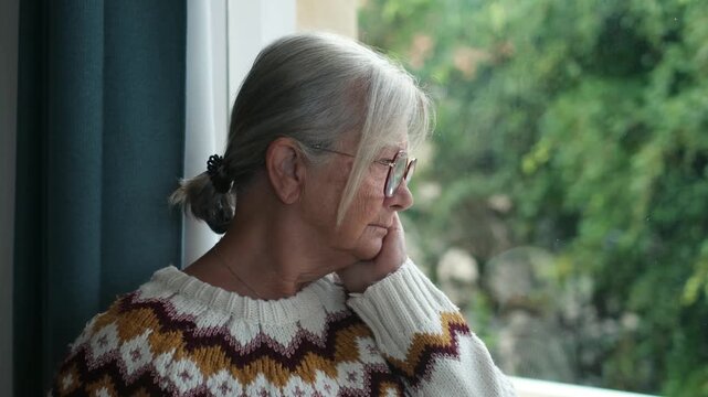 Pensive senior woman with grey hair and glasses looking out the window, feeling lonely and melancholic. She is resting her head on her hand, lost in thought and memories while at home