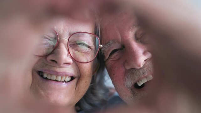 Senior couple form a heart with their hands, then lower it while laughing at the camera. Close-up focus reveals smiling elderly faces, expressing joy, affection and lasting indoor romance.