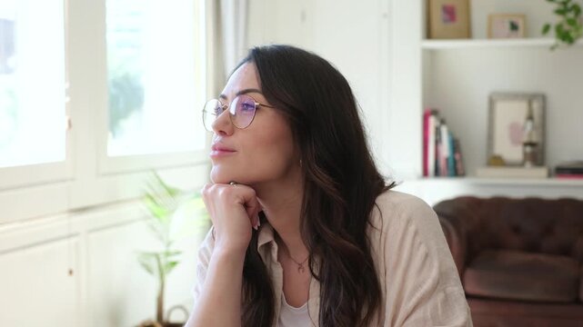 Woman rests her chin on her fist and gazes toward window light at home. Her quiet pose conveys thinking, daydreaming, personal reflection, and a calm pause in a bright living room.
