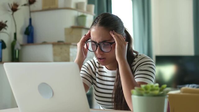 Woman pauses typing on her laptop and rubs both temples in pain at a home desk. The stressed worker shows headache, eyestrain and mental fatigue during demanding computer work in her living room.