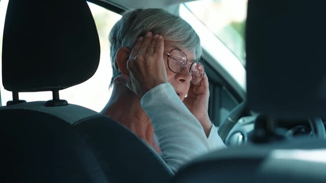 Senior woman presses both hands to her temples and grimaces from headache pain in the driver's seat. Her distressed expression illustrates migraine symptoms, driver fatigue, health concerns, and road.