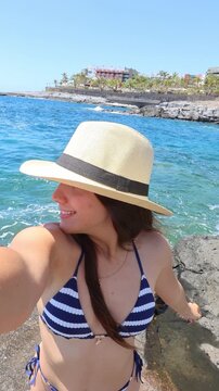 Beautiful young woman wearing a bikini and a straw hat taking a selfie video with her smartphone on a rocky beach, smiling and enjoying her summer vacation on a sunny day by the ocean