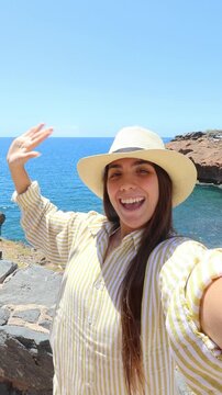Woman films a selfie, waves and makes a peace sign on a sunny coastal cliff. Her handheld travel vlog shares a cheerful ocean view, rocky shoreline and summer vacation atmosphere with viewers.