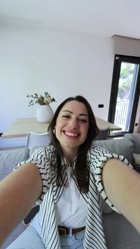 Woman laughs, winks and tilts her head while filming a selfie from her sofa at home. Handheld POV highlights playful facial expressions, direct eye contact and lively personal vlog content.