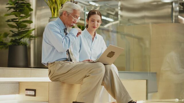 Young Thai woman with laptop smiles at thoughtful senior man