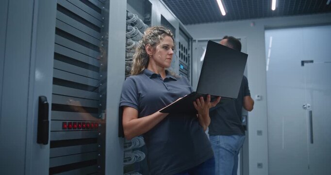 Female Server Room Specialist Monitors Server Analytics on Laptop while Colleague Inspects Nearby Racks, Showing Coordinated Infrastructure Oversight, Performance Monitoring, Diagnostics, and IT Work.