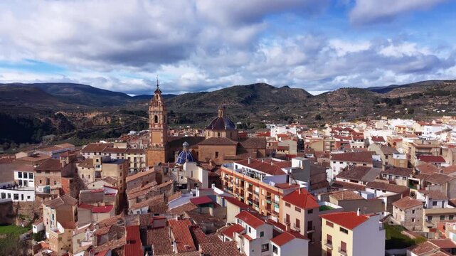 Elevated view of Chelva village in Spain with church bell tower, blue tiled domes, terracotta rooftops and rolling hills under cloudy sky