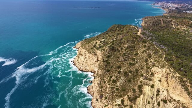 Turquoise sea waves breaking against a rugged limestone coastal cliff covered with green scrub and pine trees, with a winding dirt path along the ridge