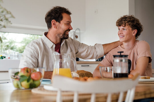 Smiling father sharing affectionate moment with teenage son at breakfast.