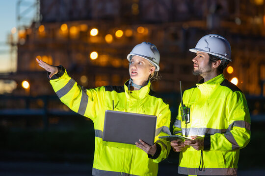 Female engineer with laptop pointing direction to colleague during night inspection at industrial plant, industrial engineers holding laptop and tablet while working at power plant facility at night