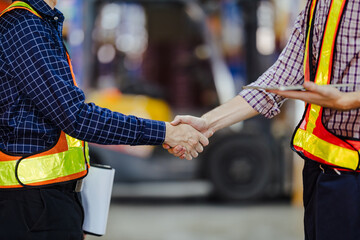 Two workers in reflective safety vests shake hands in a warehouse, symbolizing partnership and agreement in logistics, with industrial equipment blurred in the background. © akarawit