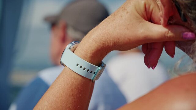 Close-up of a woman's wrist with a smartwatch on a light blue strap and pink nails, a man in a cap blurred behind, French Riviera, France.